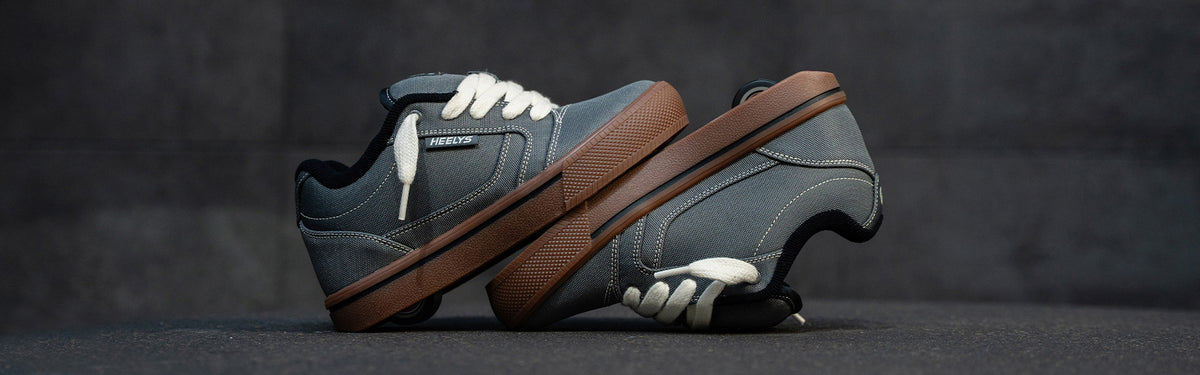 A pair of grey Pro 25 Heelys with white laces, and a thick brown sole, shown against a dark gray textured background.