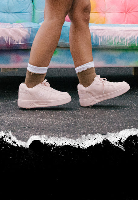 How To Heely. Person wearing light pink Heelys roller shoes glides with a colourful sofa in the background